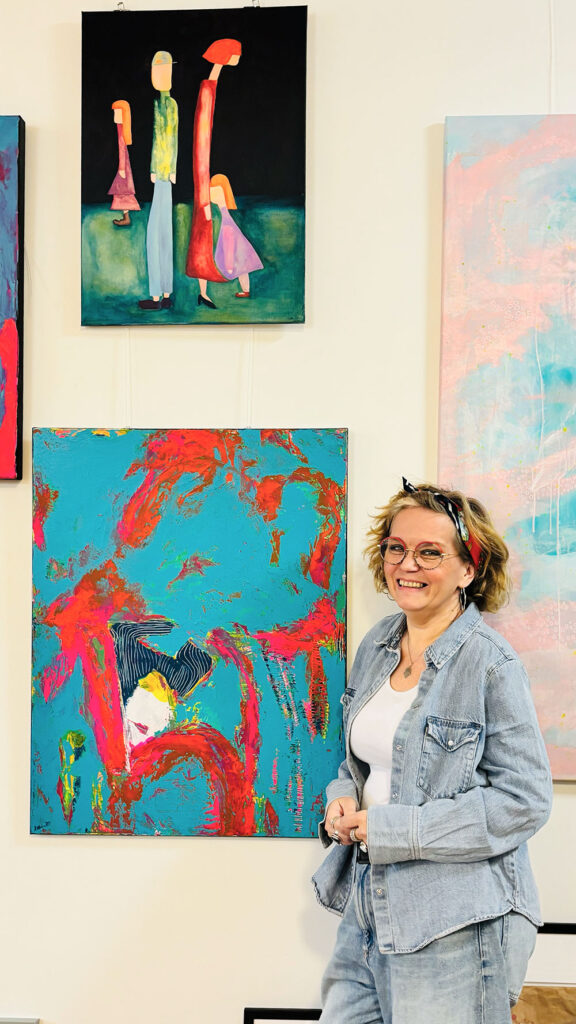 Freundlich lächelnde Künstlerin Patricia Kelm steht vor Ihren Gemälden.